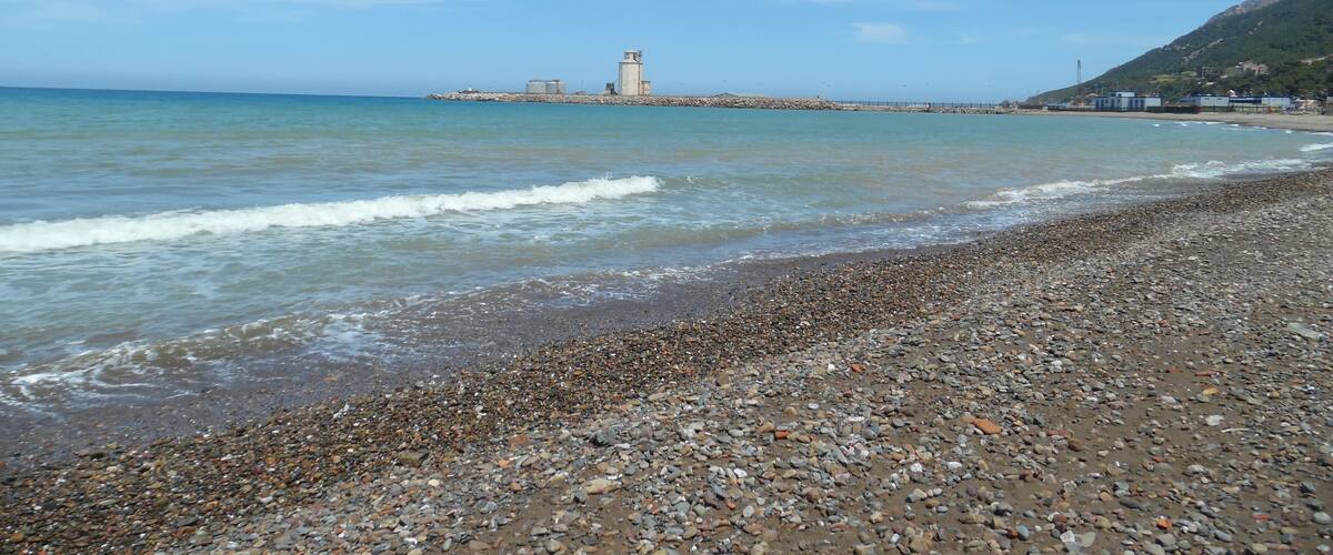 A pebbly beach with gentle waves, a distant lighthouse, and a hill with greenery.