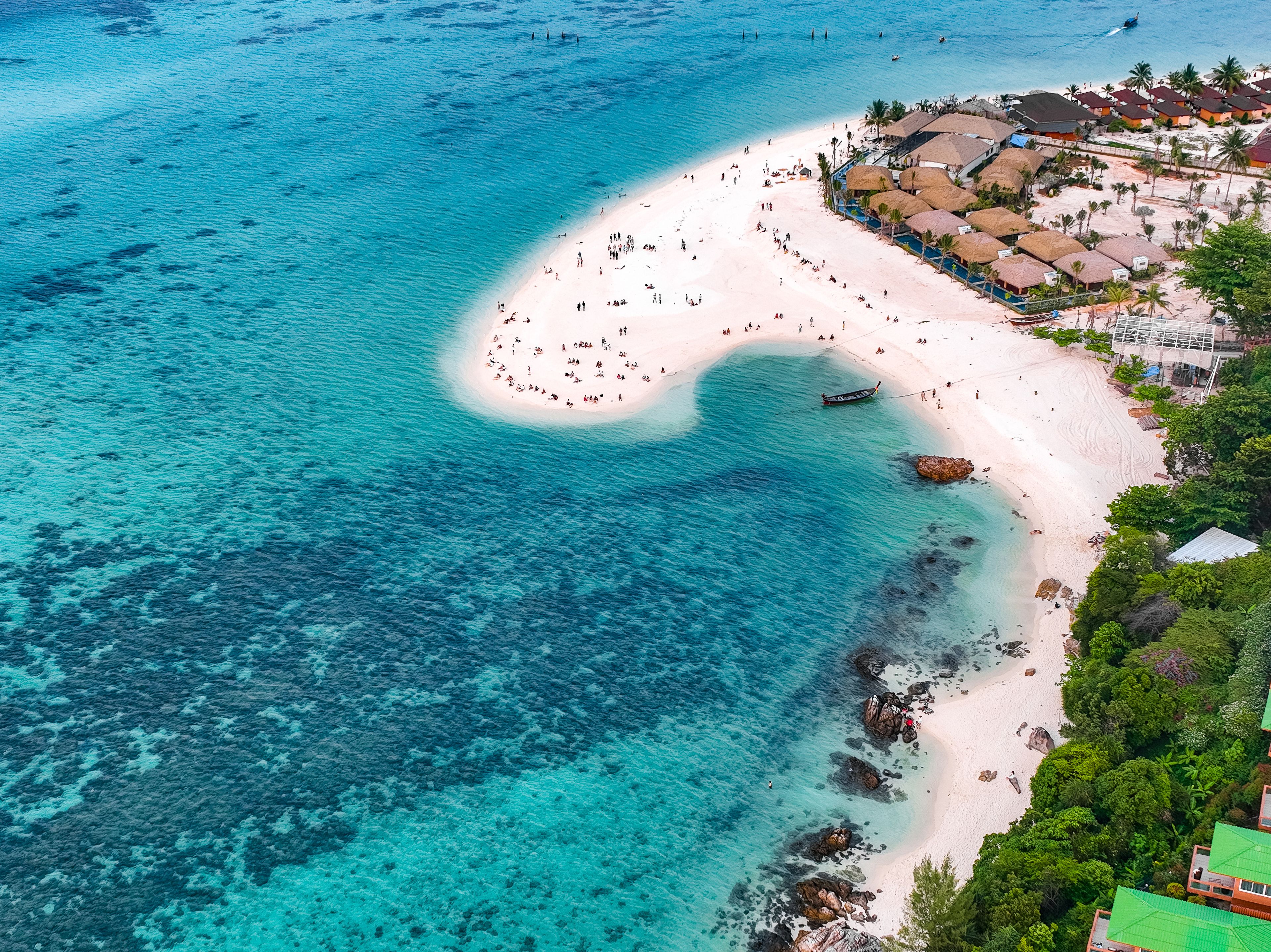 Vista aérea de uma praia com águas turquesa cristalinas, faixa de areia e um conjunto de edifícios com telhados de palha.