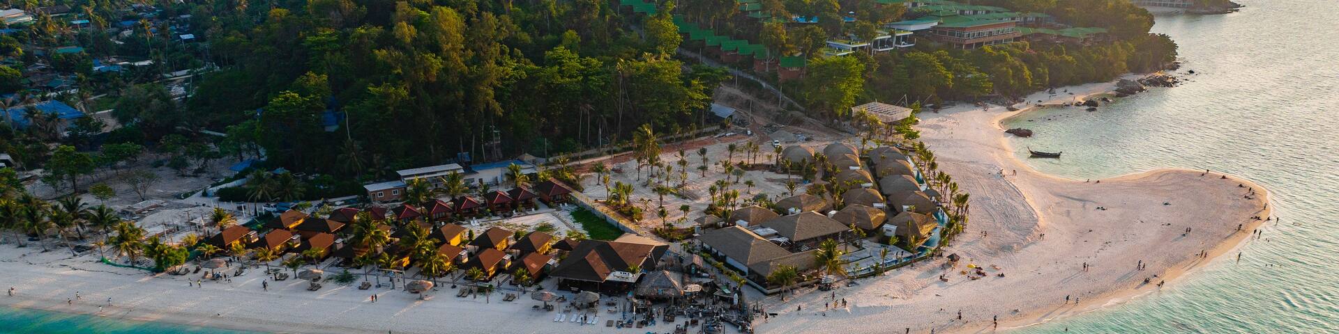 An aerial view of a coastal resort with a sandy beach, thatched-roof structures, and lush greenery.