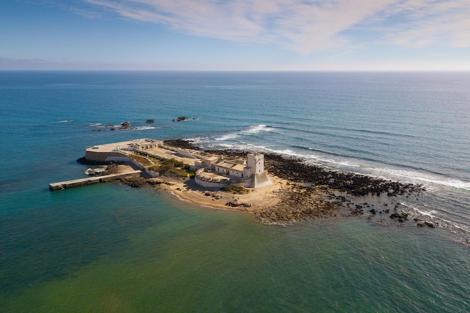 An aerial view of a coastal fortification with a tower, surrounded by the ocean and rocky shores.