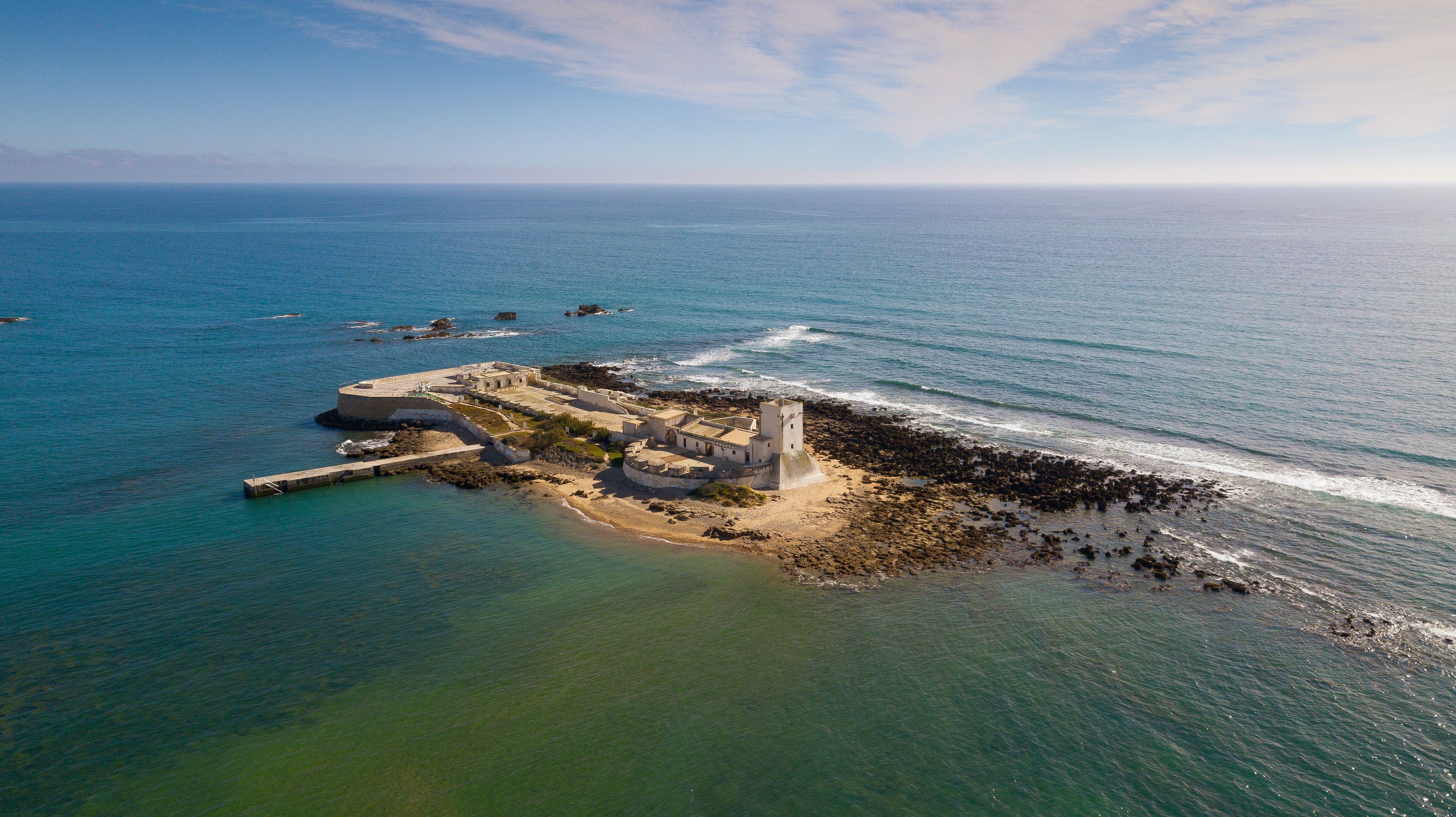 An aerial view of a coastal fortification with a tower, surrounded by the ocean and rocky shores.