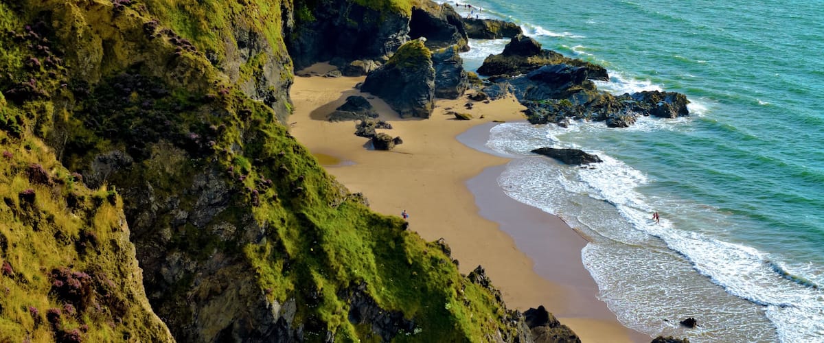 A coastal landscape with a sandy beach, rocky cliffs, and a grassy hill.