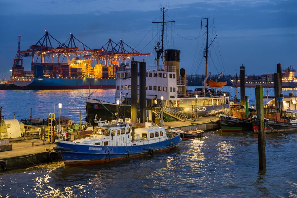 A harbor at dusk with a large cargo ship, a smaller passenger boat, and a docked ship with cranes.
