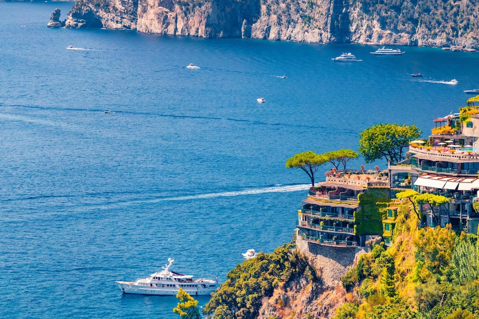 A coastal scene with a large body of water, several boats, and a cliffside building with terraces.