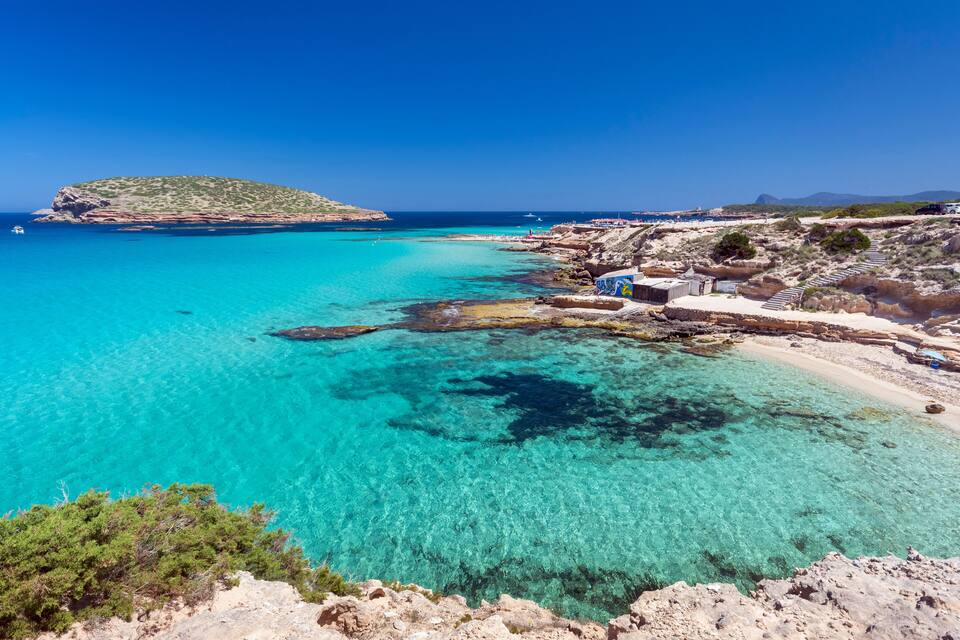 Eine Küstenlandschaft mit klarem türkisblauem Wasser, felsigen Ufern und einer kleinen Insel in der Ferne.