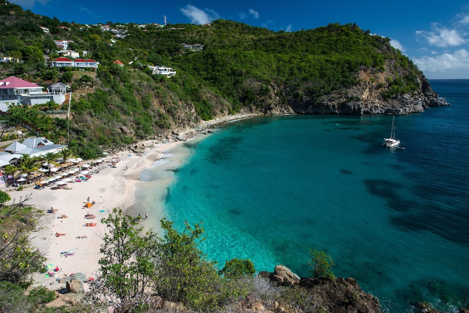 Uma praia turquesa com águas cristalinas, um veleiro e casas numa encosta.