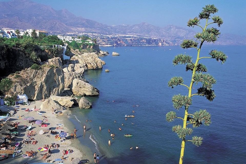 A coastal beach with rocky cliffs, clear blue sea, and a mountainous backdrop.
