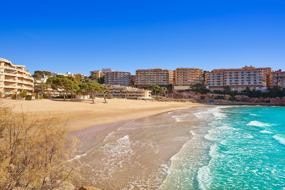 Une plage aux eaux turquoise et limpides, avec un rivage sablonneux et des immeubles résidentiels en arrière-plan.