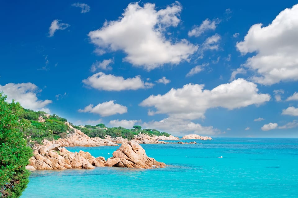 A coastal landscape with clear turquoise waters, rocky formations, and lush greenery under a bright blue sky with scattered clouds.