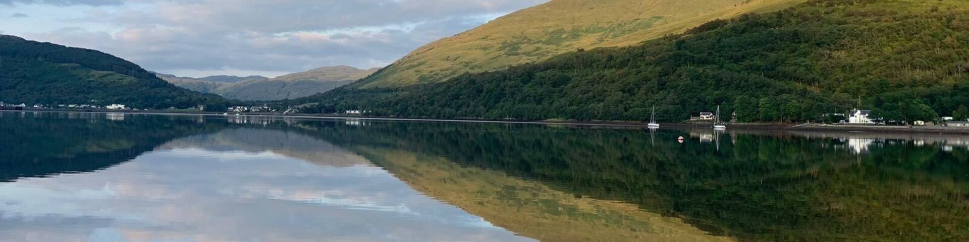 Beautiful view of Loch Long in Ardgartan, Scotland .
Clear blue water filled with pebbles and the reflection of the mountains and hills .