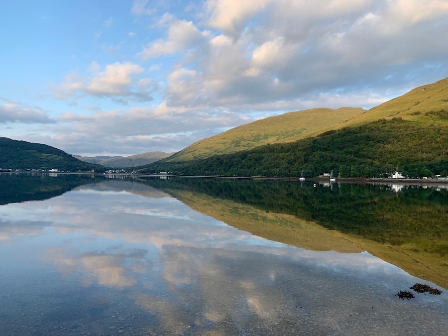 Beautiful view of Loch Long in Ardgartan, Scotland .
Clear blue water filled with pebbles and the reflection of the mountains and hills .