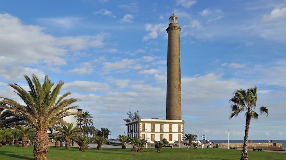 Een hoge toren met een vuurtoren, omgeven door palmbomen en een gebouw.