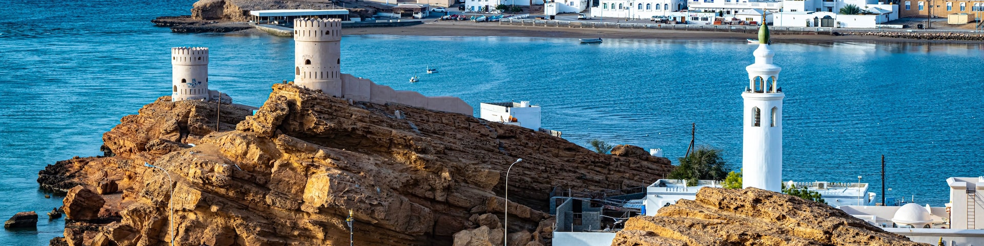 A coastal town with white buildings, a lighthouse, rocky outcrops, and boats in the harbor.