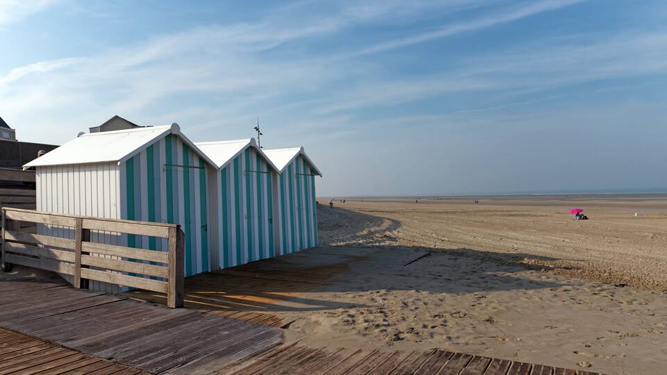 A wooden pier leading to beach huts with striped awnings, a sandy beach, and a clear sky.