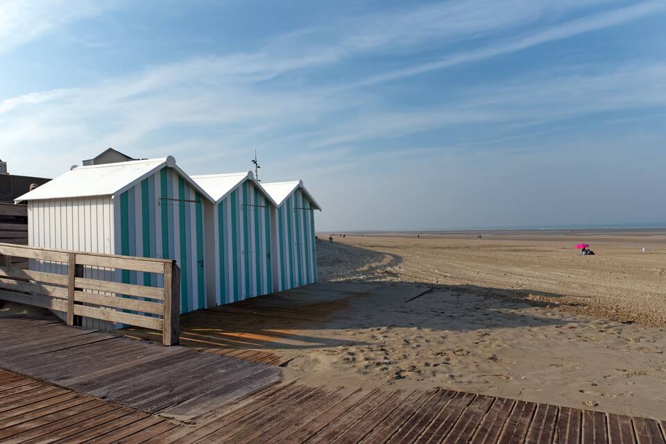 A wooden pier leading to beach huts with striped awnings, a sandy beach, and a clear sky.