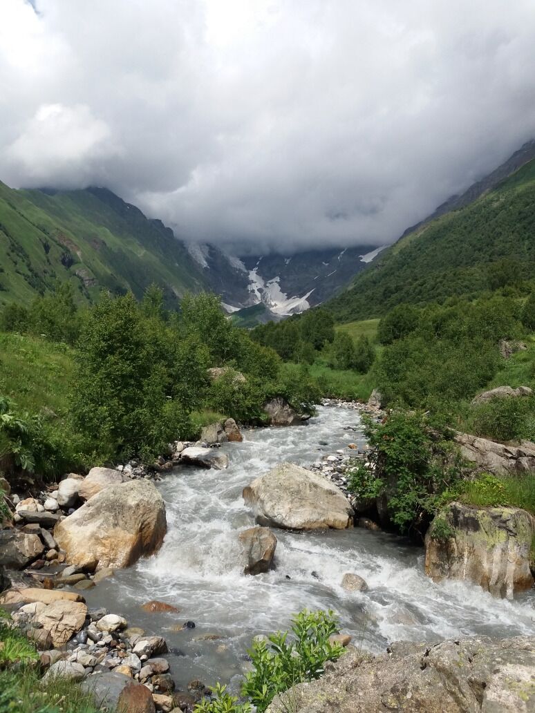 A fast-flowing river with large rocks, surrounded by lush green hills and a cloudy sky.