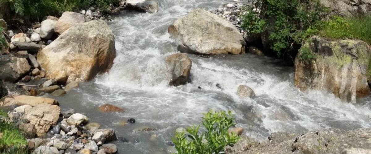 A fast-flowing river with large rocks, surrounded by lush green hills and a cloudy sky.