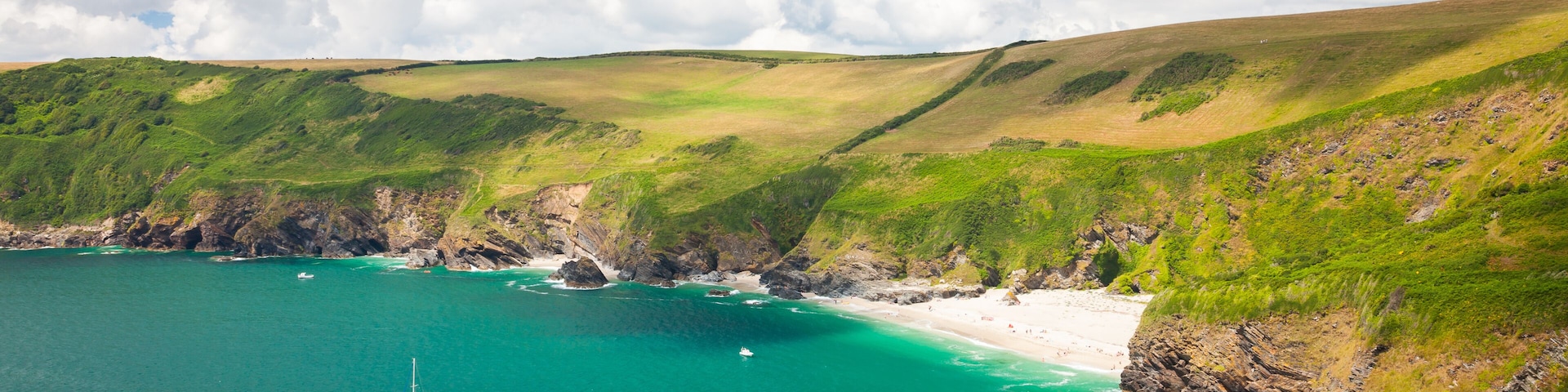 A coastal landscape with a clear blue sea, a sandy beach, and green hills.