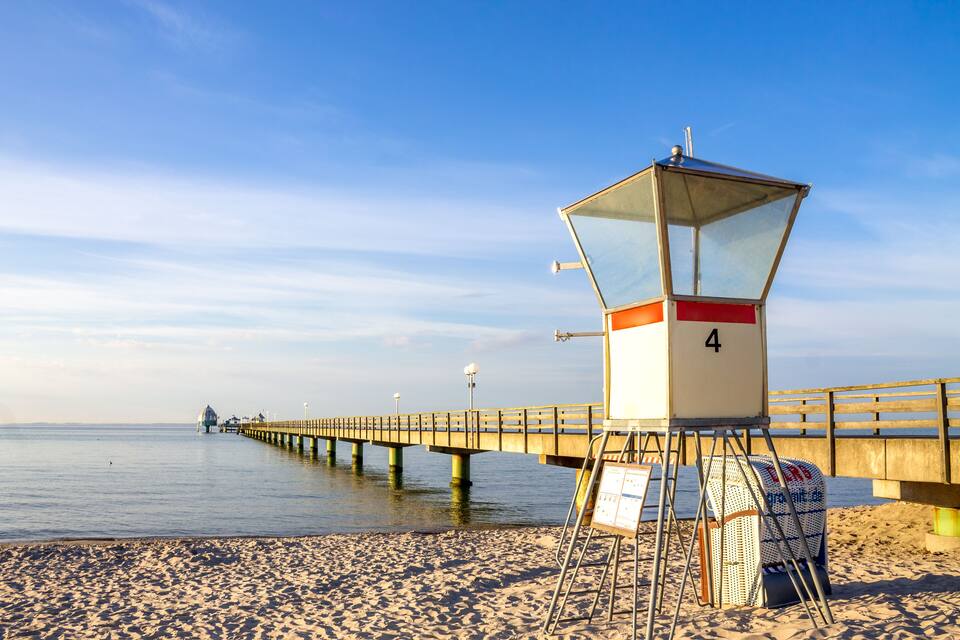 Ein Rettungsschwimmturm am sandigen Strand, mit einer hölzernen Mole, die ins Wasser ragt.