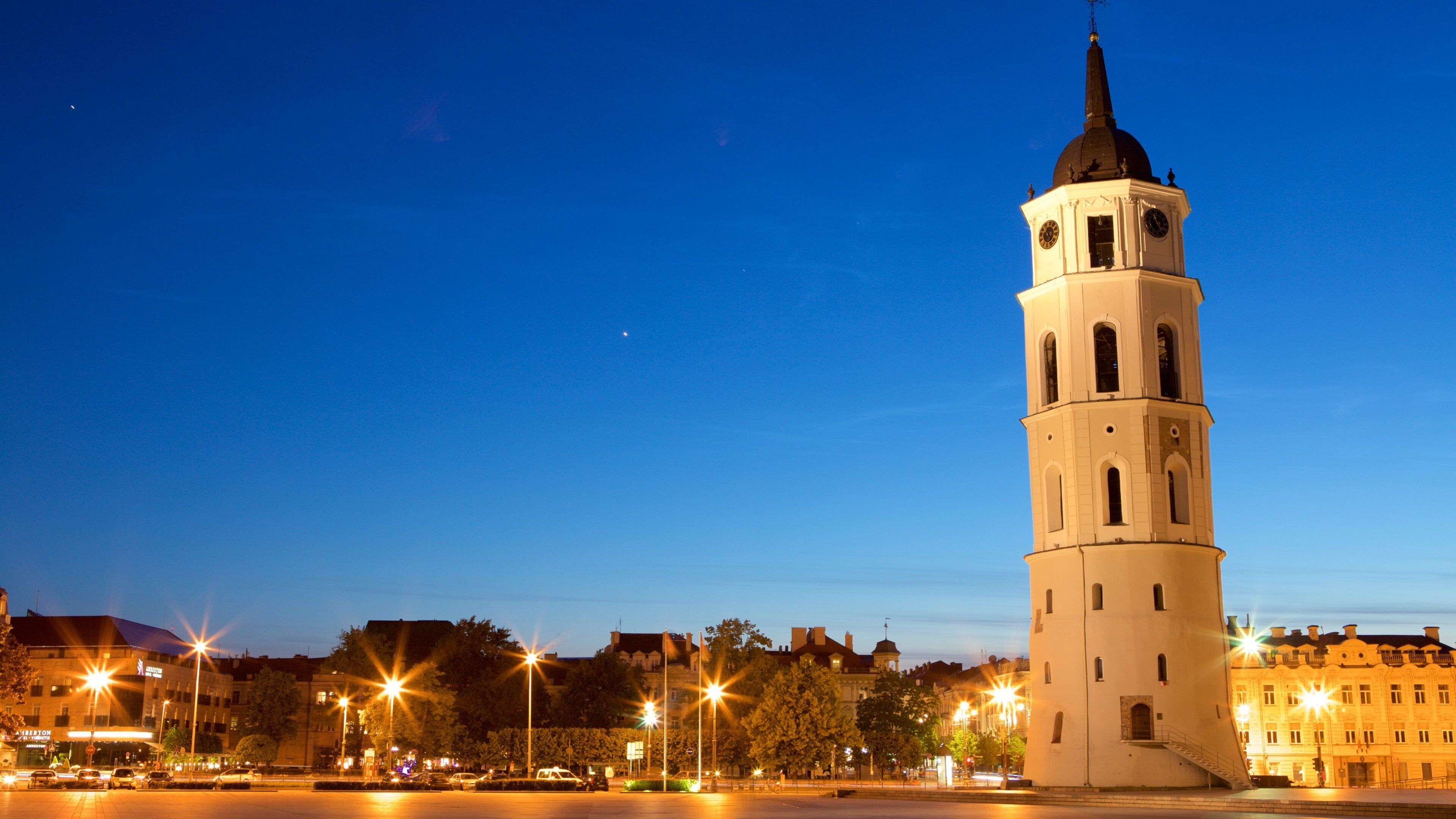 Une haute tour surmontée d’une horloge, entourée des lumières de la ville et des bâtiments.