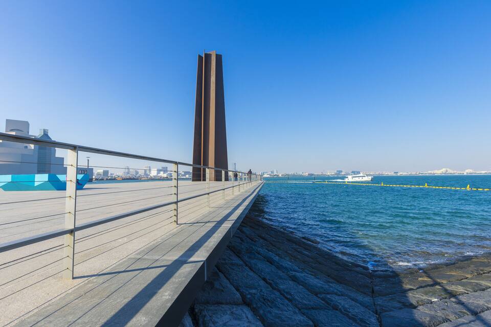 Un passeig marítim amb una torre alta i moderna, un moll de pedra i una silueta urbana llunyana.