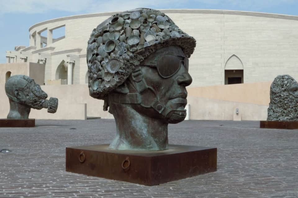 A sculpture of a soldier's head with a helmet made of metal plates, set against a building with arches.