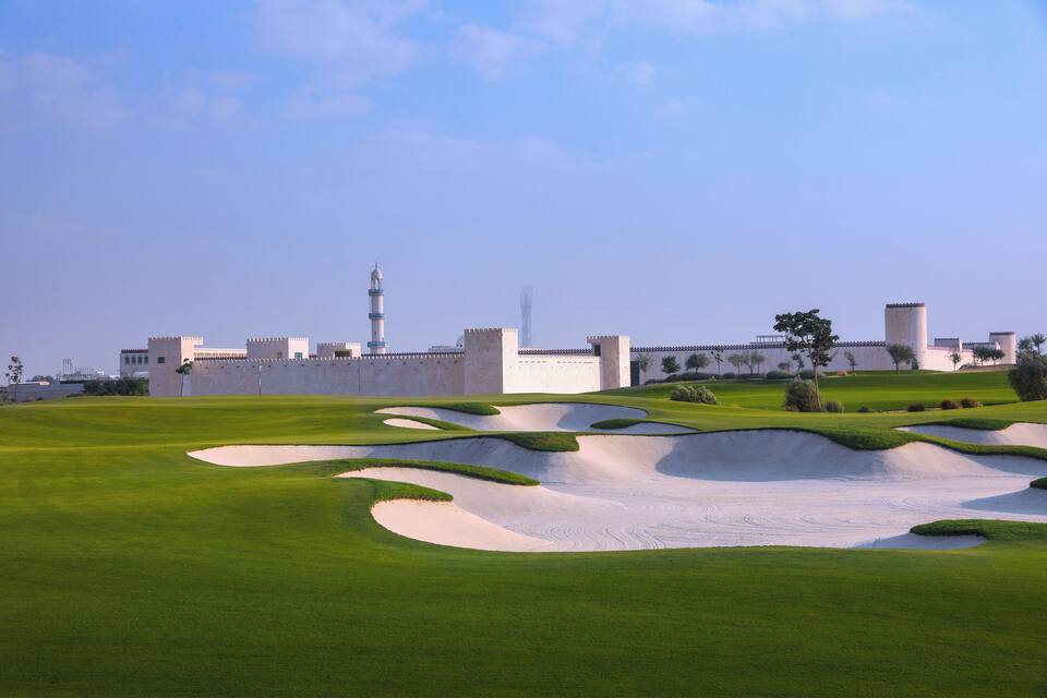 A golf course with sand bunkers and a large building with a minaret in the background.