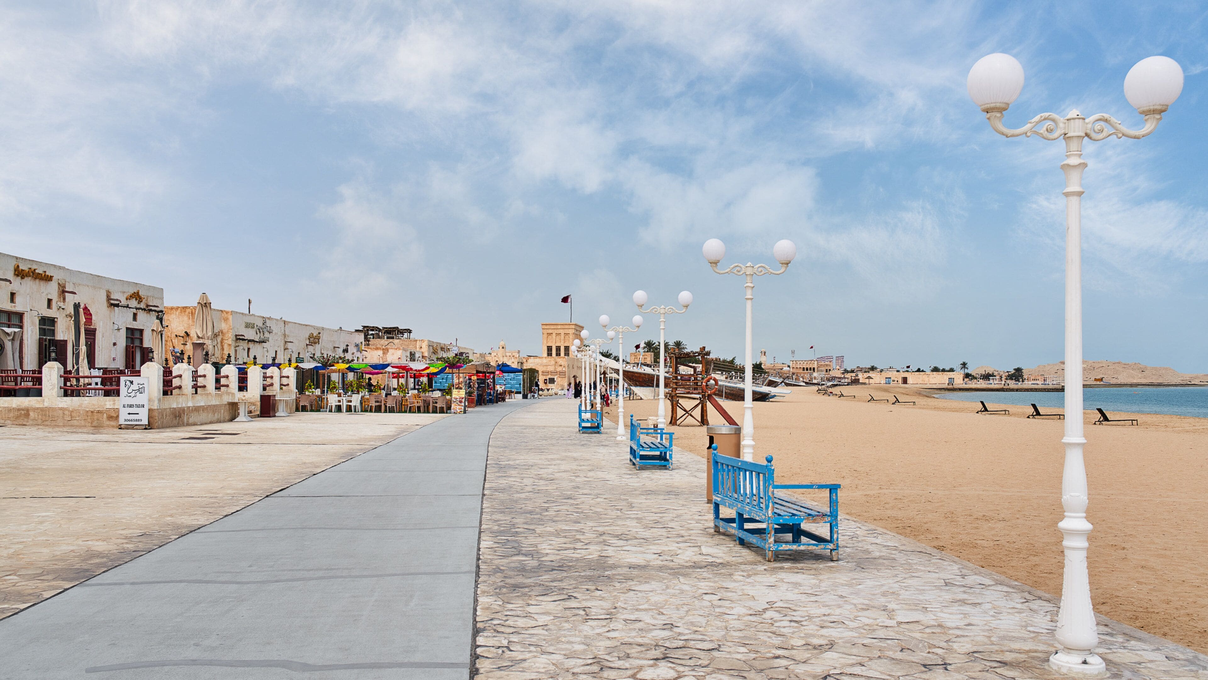A seaside promenade with a sandy beach, a row of blue benches, and a row of white street lamps.