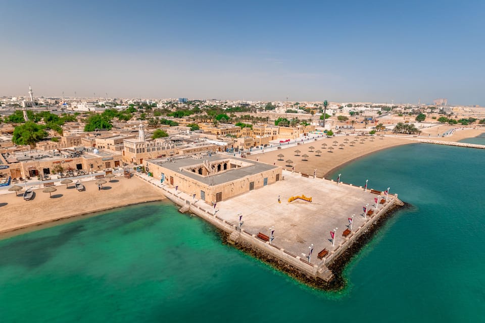 An aerial view of a coastal city with a prominent fort, a sandy beach, and a clear blue sea.