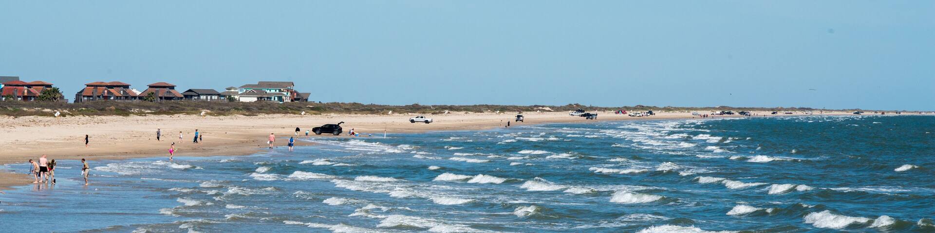 A beach with waves, people, and houses in the background.
