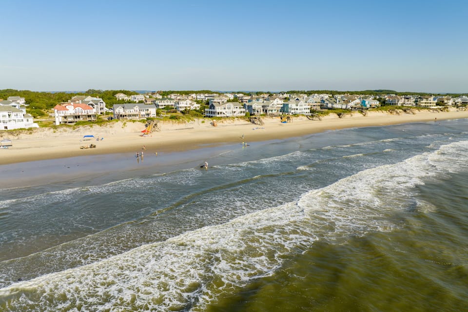 A coastal beach with houses along the shore and waves crashing onto the sand.