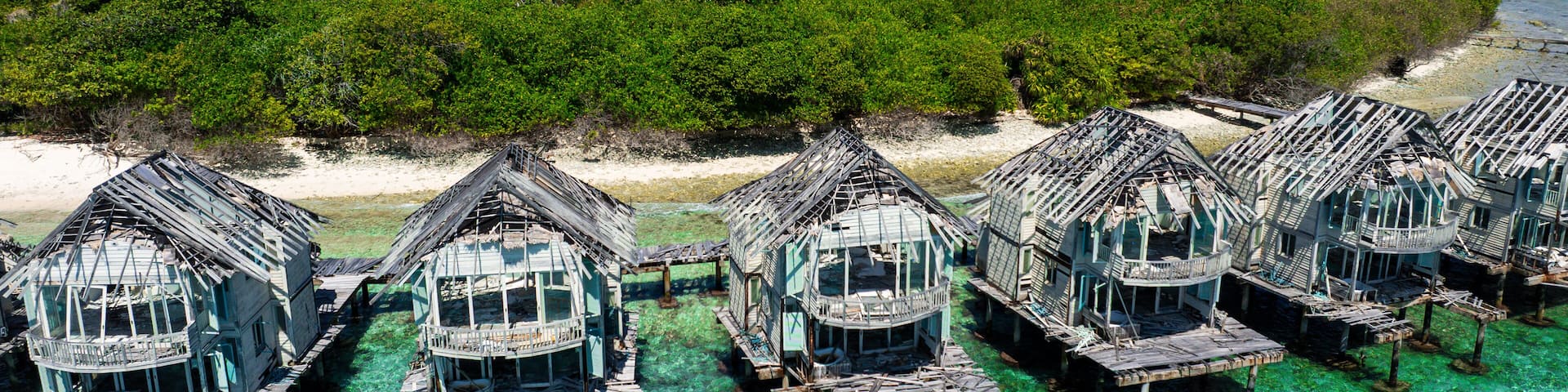 Aerial view of weathered overwater bungalows stand as silent sentinels against the azure sea, contrasting with the vibrant green island, Dhidhdhoo, Haa Alifu Atoll, Maldives.