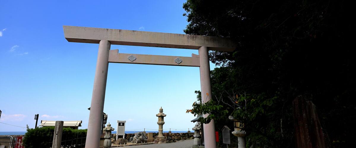 A traditional Japanese torii gate at the entrance of a shrine, with a stone lantern and a pathway leading to the sea.