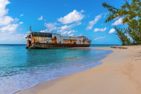 Ein verrostetes Schiffswrack am sandigen Strand, mit klarem türkisblauem Wasser und wolkenlosem, blauem Himmel.