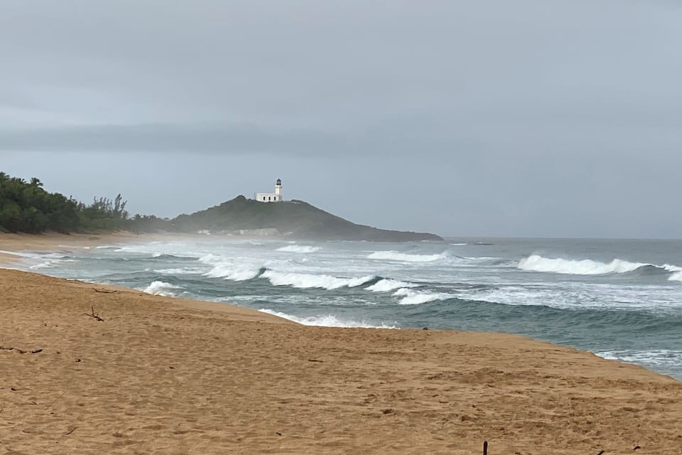 A beach with waves crashing onto the shore, a lighthouse on a small hill, and a cloudy sky.