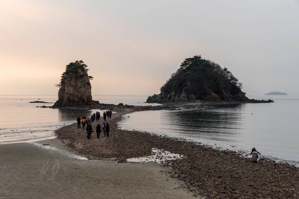Una costa rocosa con un grupo de personas caminando a lo largo de ella, dos formaciones rocosas destacadas y agua tranquila al fondo.