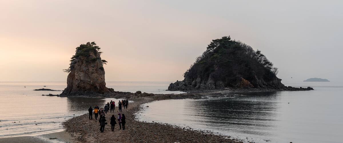 A rocky shoreline with a group of people walking along it, two prominent rock formations, and calm water in the background.