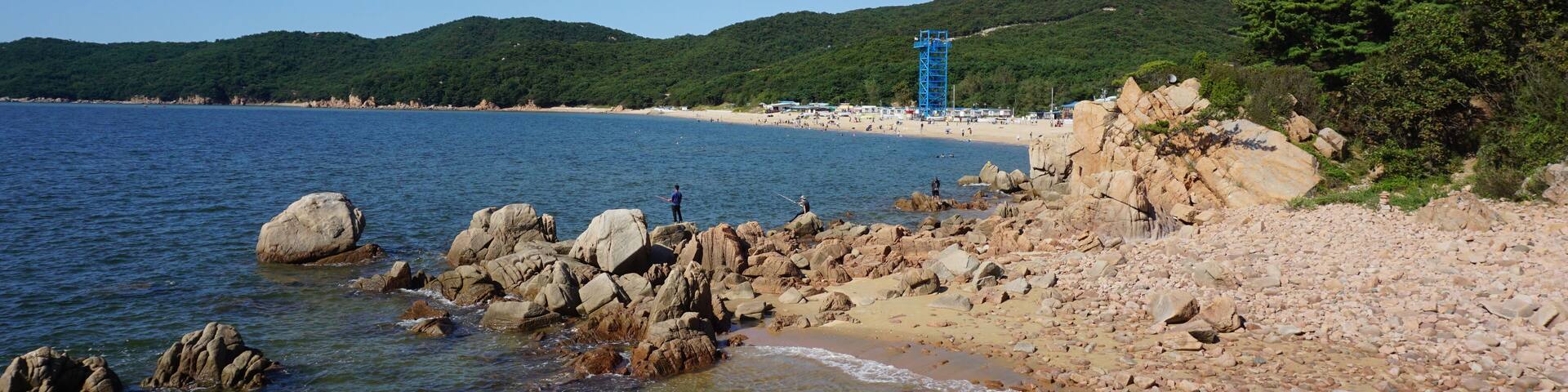 Una playa rocosa con agua cristalina, una torre de salvavidas azul y colinas verdes al fondo.