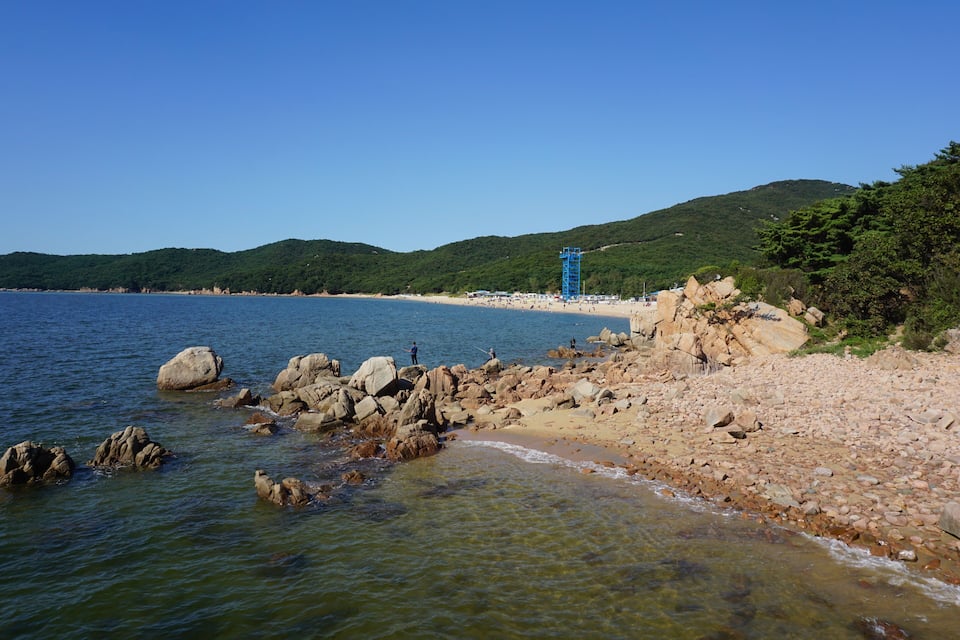A rocky beach with clear water, a blue lifeguard tower, and green hills in the background.