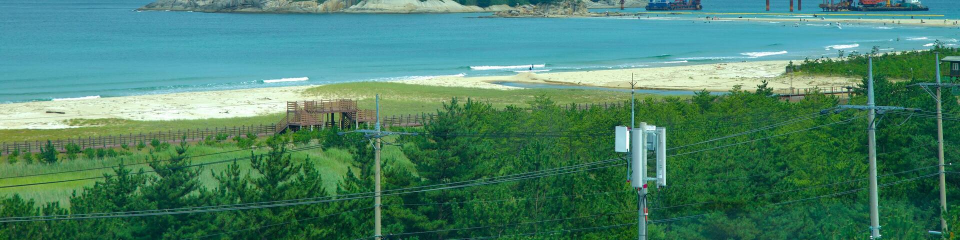Un paisaje costero con una pequeña isla, una playa de arena y estructuras industriales a lo lejos.