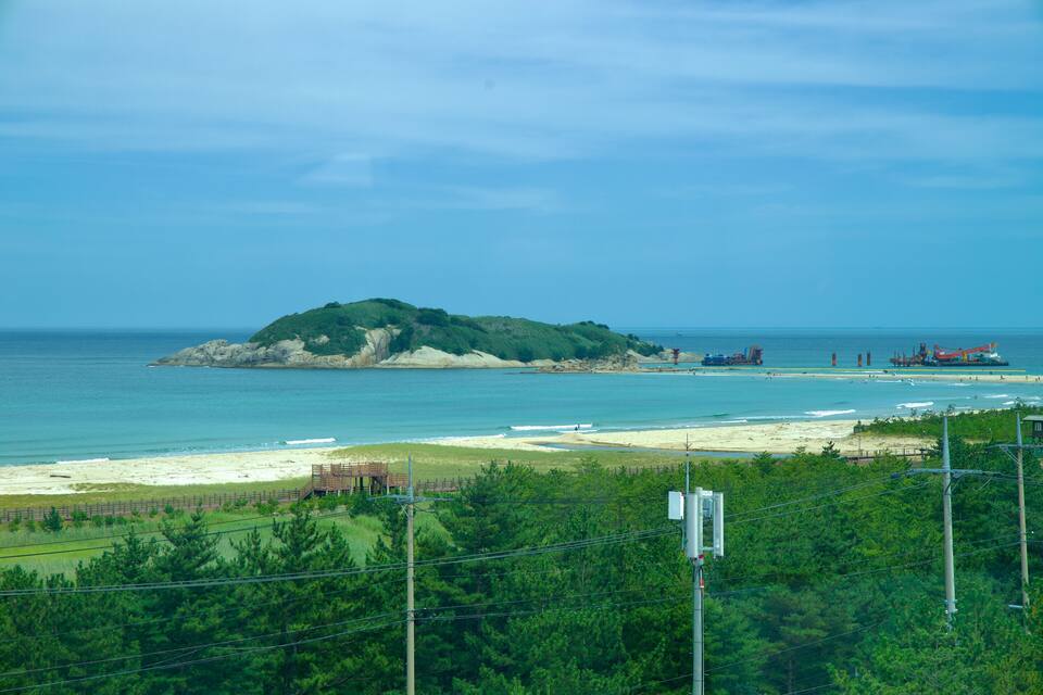 Paisagem costeira com uma pequena ilha, uma praia de areia e estruturas industriais ao longe.