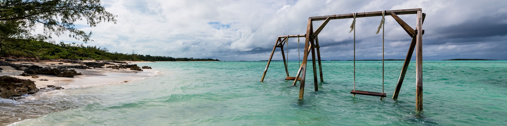 Ein hölzernes SchaukelgerĂŒst im klaren tĂŒrkisblauen Wasser, ein Sandstrand und eine von BĂ€umen gesĂ€umte KĂŒstenlinie.