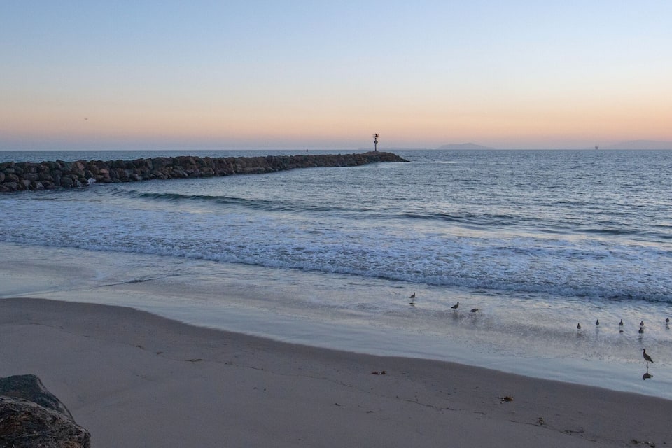 Uma praia com ondas, um paredão rochoso e um pôr do sol.