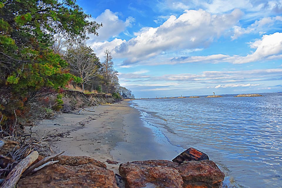 A beach with rocky outcrops, trees, and calm water under a partly cloudy sky.