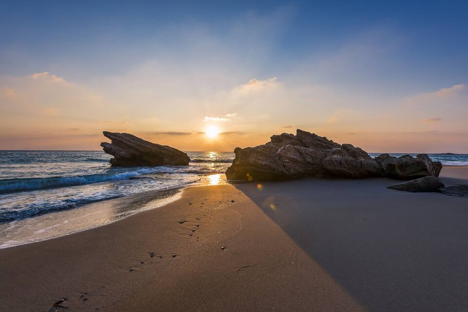 A beach with large rocks and the sun setting over the ocean.