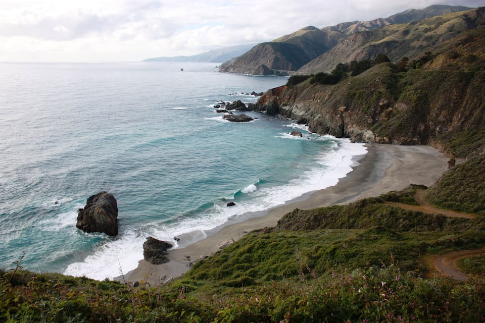 A coastal landscape with a beach, rocky shoreline, and hills in the background.
