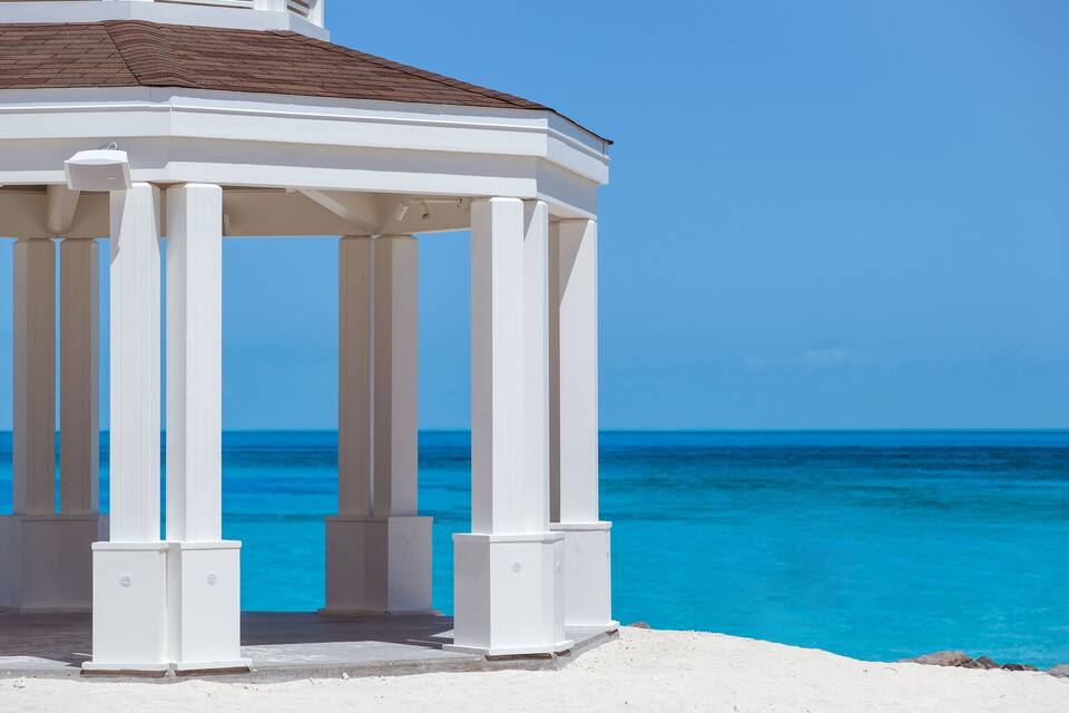 Ein weißes, säulengeträgtes Gartenhaus mit Blick auf ein türkisblaues Meer.