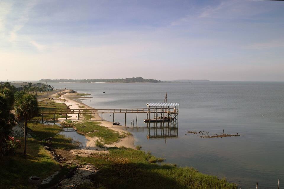Un lago tranquilo con un muelle de madera que se adentra en el agua, rodeado de vegetación y una orilla lejana.