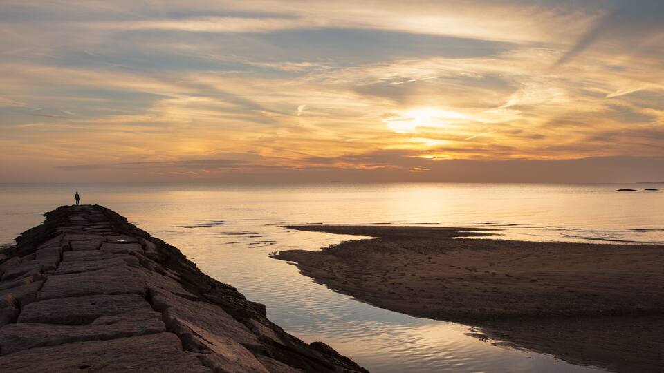 A stone pier extending into calm waters at sunset with a person standing at the end.