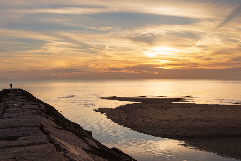 Un muelle de piedra se adentra en aguas tranquilas al atardecer, con una persona de pie al final.